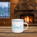 White mug with 'Colorado' on a wooden table in front of a stone fireplace with a snowy mountain view.