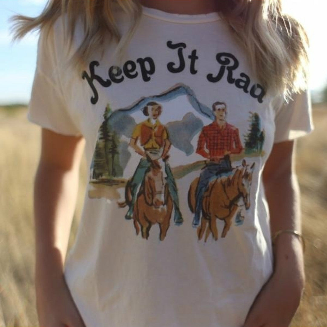Person wearing a t-shirt with a graphic of two people on horses and the text 'Keep It Rad' in a field.