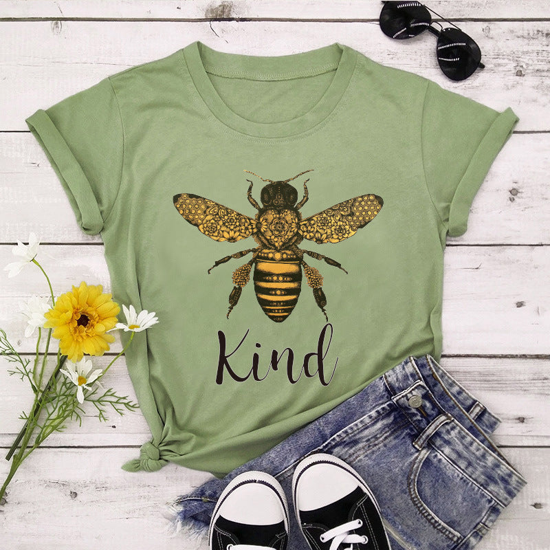 Green t-shirt with bee graphic and 'Kind' text on a wooden surface with flowers and jeans.