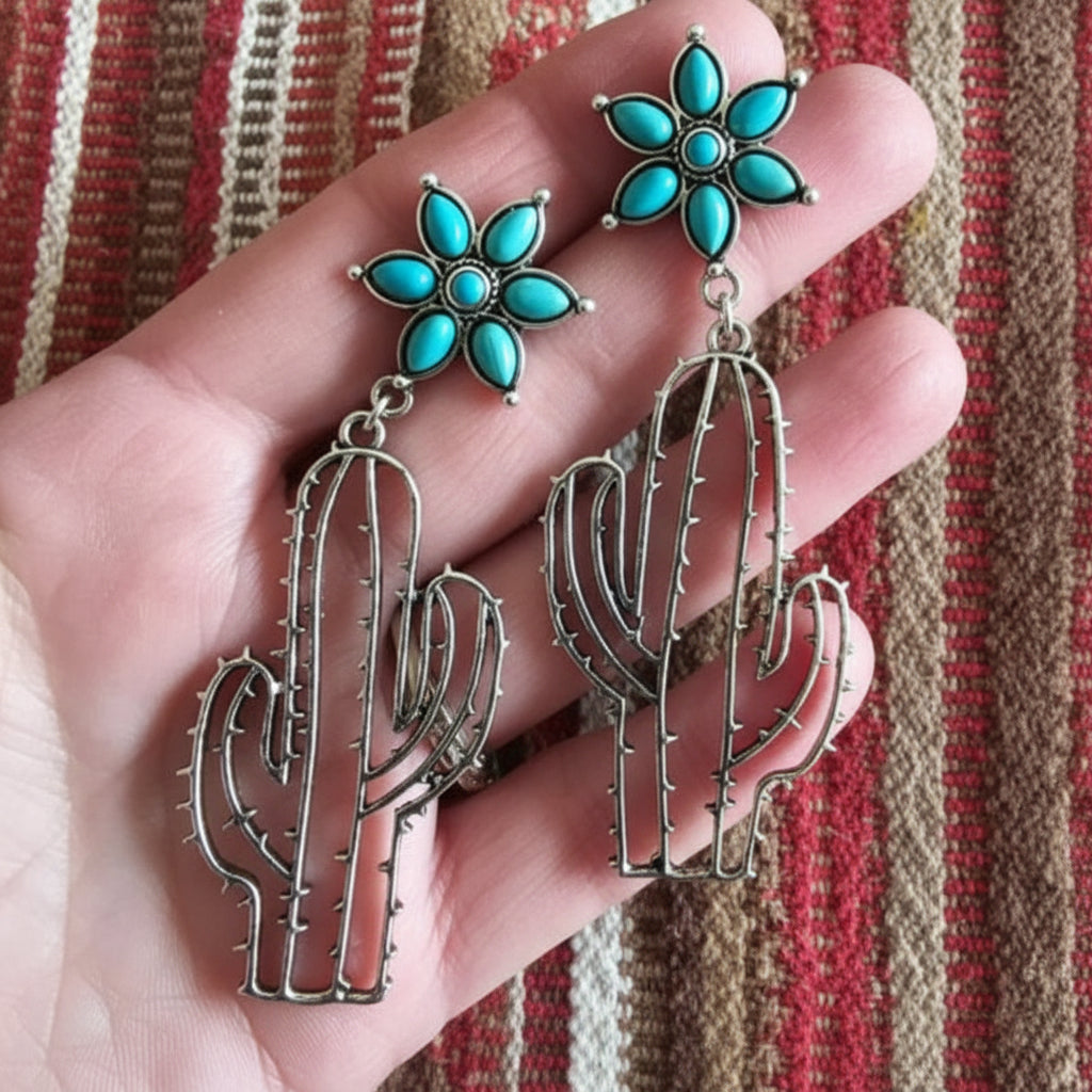Cactus-shaped earrings with turquoise stones held in a hand against a striped fabric background.